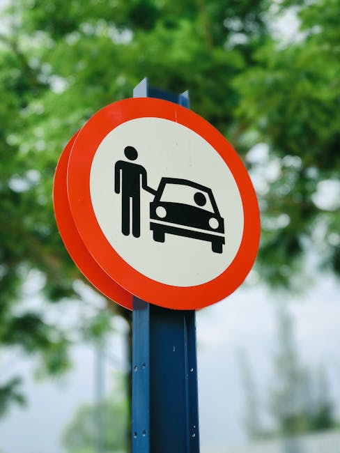 Close-up of a circular road sign with a white background and a bold red border, featuring an icon of a person standing next to a car, indicating parking restrictions or prohibition. The sign is mounted on a blue pole situated outdoors, with blurred green foliage and trees in the background. The image is relevant to house removals and moving services provided by Man with Van Becontree Heath, highlighting road regulation awareness during furniture transport and home relocation processes.