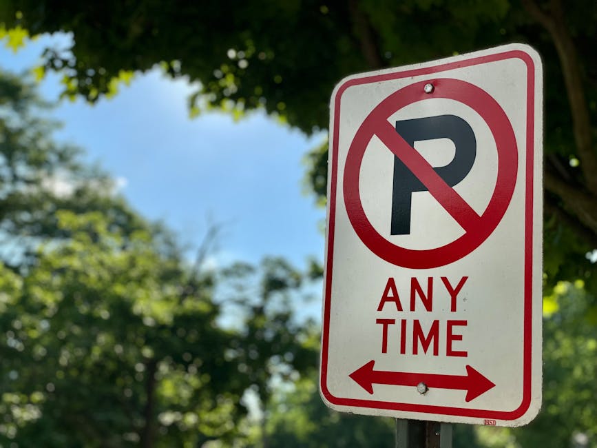 A rectangular traffic sign mounted on a metal pole outdoors, indicating no parking at any time with a large black 'P' crossed out by a red circle and line. Below the symbol, the sign reads 'ANY TIME' in red capital letters. The background includes a clear blue sky partially obscured by green leafy trees, suggesting a residential or urban area. The sign is situated near the edge of a pavement or sidewalk, with the tree foliage providing shade and natural light highlighting the sign’s surface. This signage is relevant to home relocation or furniture transport activities managed by Man with Van Becontree Heath, ensuring compliance with local parking regulations during moving or packing and moving processes.