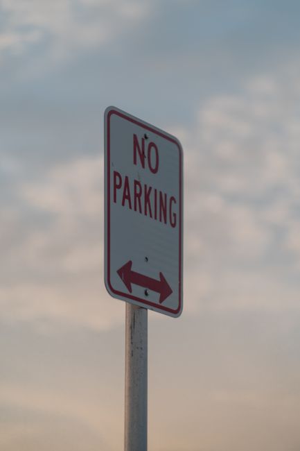 Close-up of a circular road sign with a white background and a bold red border, featuring an icon of a person standing next to a car, indicating parking restrictions or prohibition. The sign is mounted on a blue pole situated outdoors, with blurred green foliage and trees in the background. The image is relevant to house removals and moving services provided by Man with Van Becontree Heath, highlighting road regulation awareness during furniture transport and home relocation processes.