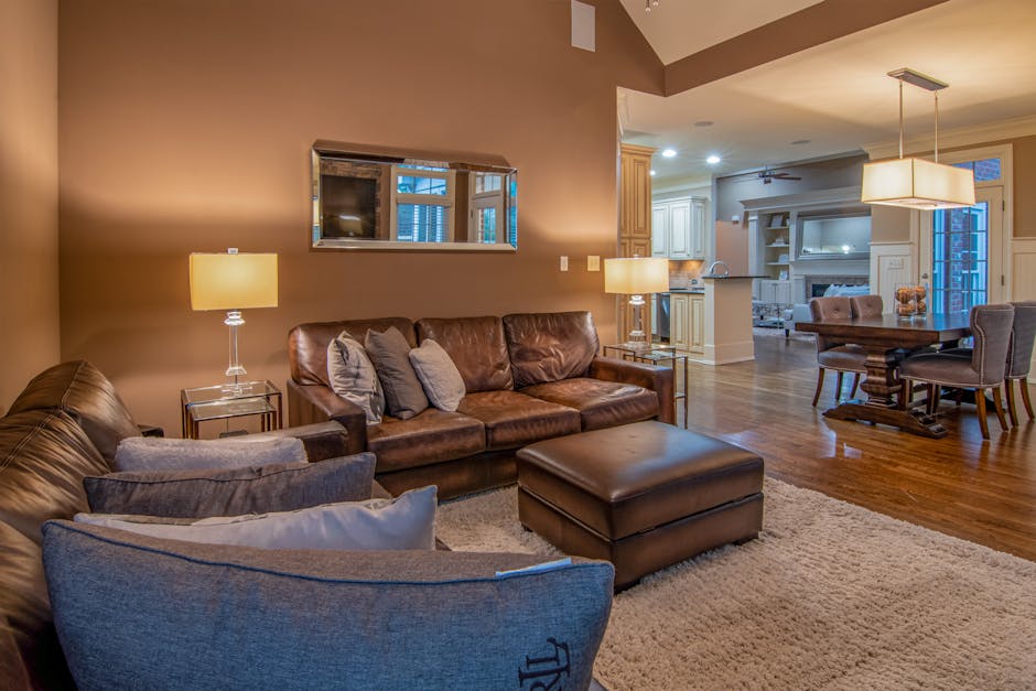 Interior view of a living room during a house removal process, showcasing a brown leather sofa with multiple grey and beige cushions, positioned against a warm beige wall with a rectangular mirror above it. Beside the sofa, there are two glass-topped side tables each illuminated by a table lamp with a beige shade. In the foreground, part of a large blue fabric armchair with a decorative cushion is visible, resting on a cream shag rug. The open-plan space extends into a dining area with a wooden table and upholstered chairs, and a modern white kitchen with cabinetry, countertops, and built-in appliances beyond. The room is well-lit with ambient lighting, and a large window and glass door in the background reveal an outdoor balcony area. This scene depicts the typical environment where furniture and household items are arranged or prepared for moving, with the emphasis on home relocation, packing, and furniture transport management by Man with Van Becontree Heath.