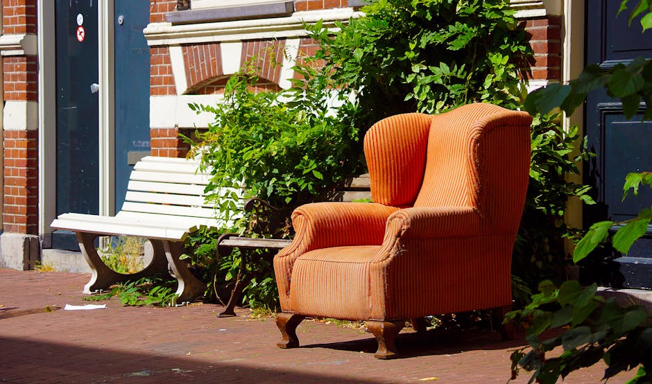 An orange upholstered armchair with a high back and curved wooden legs is positioned outside on a brick pavement, next to a white wooden bench with black metal armrests partially covered by green foliage. Behind the armchair is a brick building with white accents around the window and door. The scene is well-lit, with sunlight casting shadows on the pavement. This setting depicts the preparation phase of home relocation or furniture transport, where furniture and household items are temporarily placed outdoors before being loaded into a vehicle by a professional removal service such as Man with Van Becontree Heath, supporting efficient packing and moving processes.