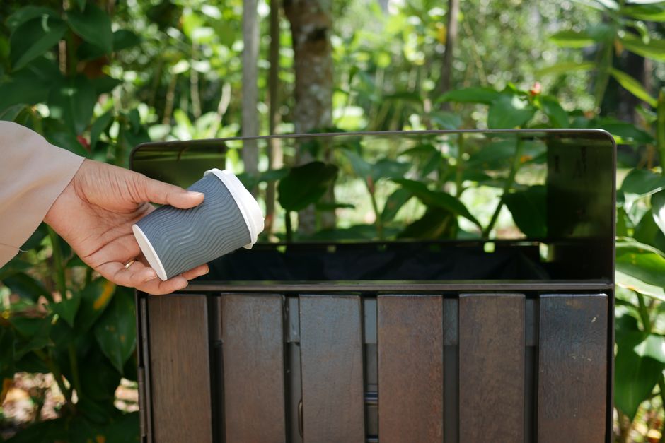 A person’s hand placing a used paper coffee cup into a metal recycling bin situated outdoors amidst green foliage and trees; the recycling bin has a rusted, dark metal body with vertical slats and a reflective lid. The scene is illuminated by natural daylight, highlighting the outdoor environment, which appears to be in a backyard or garden area, with the focus on waste disposal during a home relocation or decluttering process. Man with Van Becontree Heath offers services related to removals and bulk waste disposal, supporting efficient home or furniture transport and packing and moving logistics.
