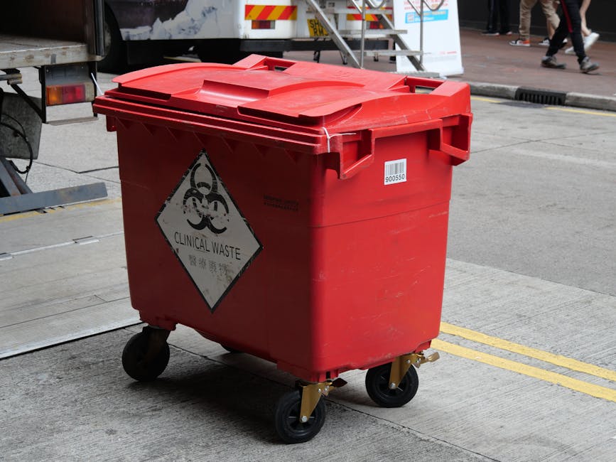 A red clinical waste bin with a hinged lid on four black caster wheels is positioned on a concrete sidewalk outside a building. The bin features a hazardous biohazard symbol and a white label with a barcode on its front. In the background, part of a vehicle with a loading ramp and a group of pedestrians walking along a paved street are visible, suggesting a busy urban environment. This scene relates to household or commercial waste disposal, which is often involved in home relocation or clearance services offered by companies like Man with Van Becontree Heath during furniture transport or packing and moving activities.