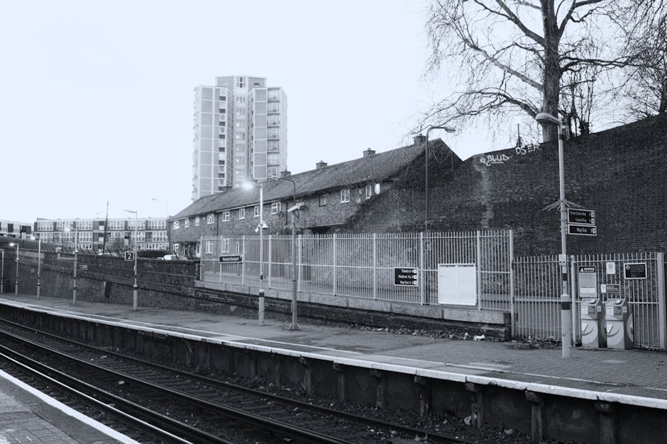 Black and white photograph of a residential area near a train station platform, with a high-rise building in the background and older low-rise houses in the midground. The platform features a metal railing, street lamps, and various signage for train services, including a timetable display. Two payphones are mounted on a metal stand at the edge of the platform. The scene includes a paved surface with a concrete curb separating it from the railway tracks, which are empty in this image. Overhead wires are visible above the tracks, and street lamps illuminate the area. A large, leafless tree stands beside the platform, with some graffiti visible on the wall behind the low-rise houses, indicating urban surroundings. The overall scene suggests a quiet moment at a commuter station in a residential district. This setting relates to house removals or furniture transport, akin to the logistics involved in home relocation or moving services provided by Man with Van Becontree Heath.
