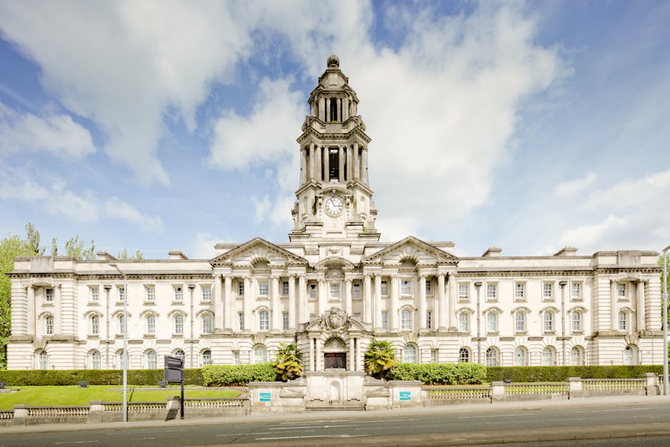 Photograph of a large, historic building with elaborate architectural details, symmetrically designed with multiple windows and a prominent clock tower at the center. The building is constructed from light-colored stone and features decorative elements such as columns and carvings. In front of the building, a well-maintained hedge lines the sidewalk, and several small potted plants are placed near the entrance. The sky above is partly cloudy with patches of blue, and the surrounding environment includes a paved street with no visible traffic or parked vehicles. The scene suggests a public or civic building, typical of London architecture. This setting may be used as background imagery for content about house removals or property-related services, including moving and furniture transport, especially in the context of planning for home relocation in Becontree Heath, where local council rules regarding permits might be relevant.