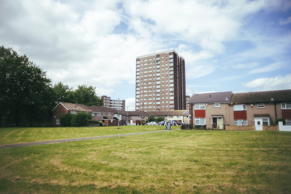 A wide view of a residential area in Becontree Heath featuring a large, open grass field in the foreground, with a paved footpath running through it. Beyond the grass, there are several small, detached houses with brick and weatherboard exteriors, some with visible front doors, windows, and garden fences. In the background, a tall multi-storey apartment building dominates the skyline, constructed with patterned brickwork and numerous windows, indicating high-density housing. The scene is illuminated by natural daylight under a partly cloudy sky, providing clear visibility of the environment. This setting illustrates a typical neighbourhood where house removals and furniture transport by Man with Van Becontree Heath could involve outdoor loading areas, with steps of packing, loading vans, and transport logistics visible in the context of home relocation procedures.