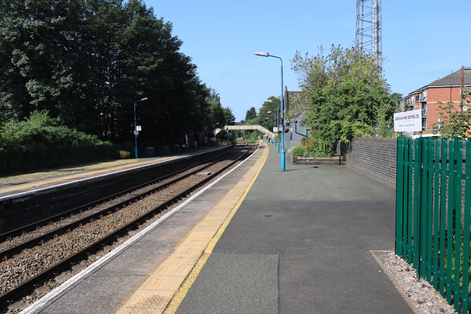 The image shows Becontree Station platform in daylight, with railway tracks running parallel along the left side and a concrete platform on the right. The platform features a yellow tactile paving strip near the edge for safety, and a series of blue-painted lamp posts providing lighting. In the background, a pedestrian bridge arches over the tracks, and there are trees and shrubs on the far side, suggesting a suburban setting. On the right, a dark brick wall and a green fence enclose a small garden area with flowering plants and foliage. The platform appears clear and ready for passengers, with no visible moving equipment or personnel, capturing a quiet moment during a typical day. For house removals or furniture transport, the scene highlights the importance of clear, accessible pathways for loading and unloading, which [COMPANY_NAME] can assist with in relocating belongings efficiently, whether inside property or onto transporting vehicles for home relocation or packing and moving processes.