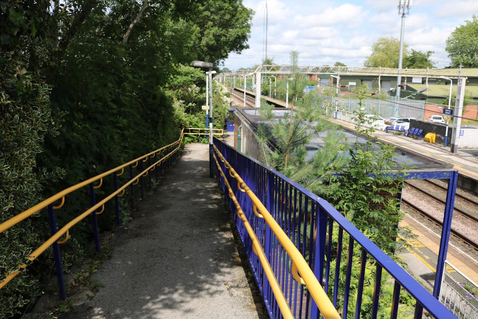 A narrow outdoor footpath with a black asphalt surface and yellow safety railings on both sides, running adjacent to a railway station platform visible in the background. The path is flanked by dense green foliage and trees on the left, providing shade, while the right side overlooks train tracks, platform area, and parking lot with a few cars. The sky above is partly cloudy with some blue sky visible. The scene appears to be part of a home relocation process, with the pathway used for moving equipment or belongings from a property towards the station for transportation, with promotional context linked to [COMPANY_NAME]’s removals services. The image demonstrates the access route used during furniture transport or packing and moving activities as part of house removals.