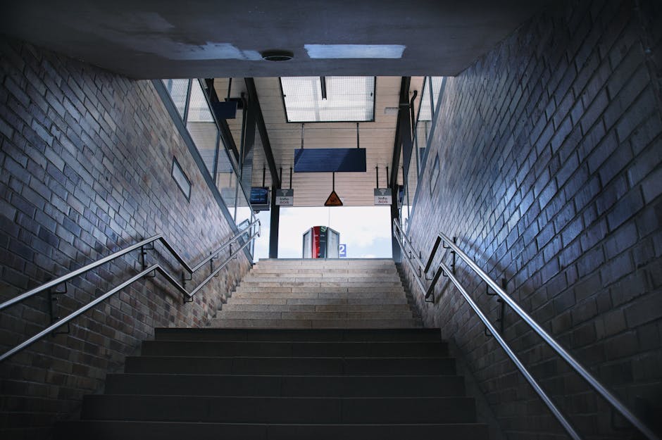 The image shows Becontree Station platform in daylight, with railway tracks running parallel along the left side and a concrete platform on the right. The platform features a yellow tactile paving strip near the edge for safety, and a series of blue-painted lamp posts providing lighting. In the background, a pedestrian bridge arches over the tracks, and there are trees and shrubs on the far side, suggesting a suburban setting. On the right, a dark brick wall and a green fence enclose a small garden area with flowering plants and foliage. The platform appears clear and ready for passengers, with no visible moving equipment or personnel, capturing a quiet moment during a typical day. For house removals or furniture transport, the scene highlights the importance of clear, accessible pathways for loading and unloading, which [COMPANY_NAME] can assist with in relocating belongings efficiently, whether inside property or onto transporting vehicles for home relocation or packing and moving processes.