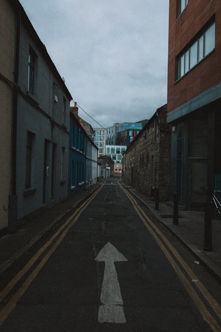 A narrow urban street scene during daytime shows a small road with an upward-pointing white arrow painted on the asphalt, flanked by old and modern buildings on either side. To the left, a row of grey, weathered terraced houses with small windows and flat roofs, while on the right, a taller, contemporary building with large glass windows and a brick facade. The street is lined with short black bollards and has double yellow lines along the edges indicating parking restrictions. The sky is overcast, casting diffuse natural light over the scene. This setting resembles a confined city lane often used during house removals or furniture transport in urban areas, which [manwithvanbecontreeheath.co.uk](https://manwithvanbecontreeheath.co.uk) services for home relocation and moving logistics. An environment suitable for vehicle parking or loading, with no visible moving equipment or furniture in sight, emphasizing the street’s role in facilitating tight-street removals and packing and moving operations.