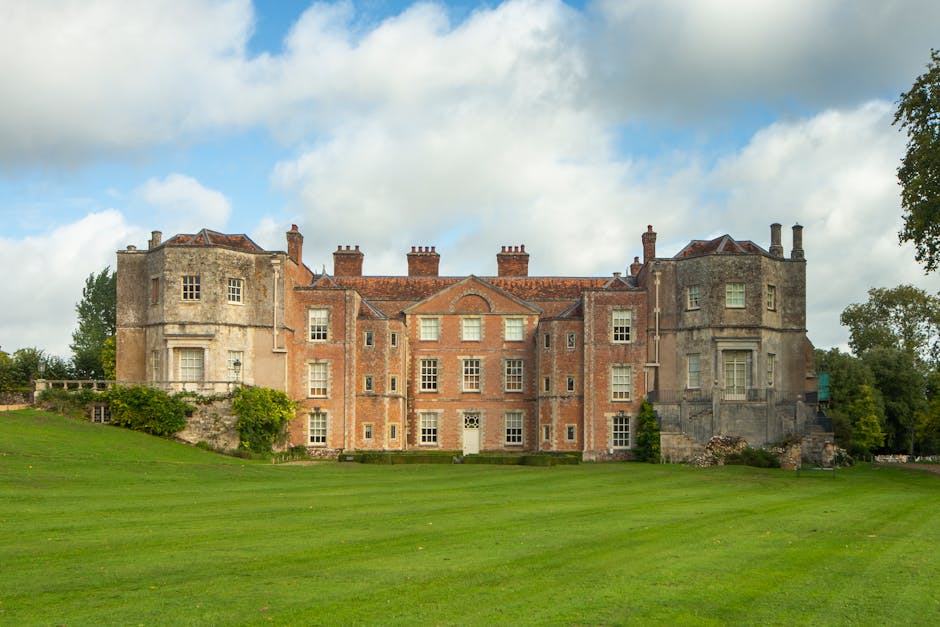 The image shows a large historic castle-style building with a stone and brick facade, multiple chimneys, and a symmetrical arrangement of windows. In the foreground, there is a well-maintained grassy lawn, with a few trees visible on the right side of the frame. The building appears to be situated at the end of a driveway or path, with the exterior walls showing signs of age and traditional architectural features such as turret-like sections on either side. The sky above is partly cloudy with patches of blue, providing natural lighting that highlights the textured surfaces of the castle's stonework. This setting is indicative of a property suitable for house removals or relocation services, and the scene emphasizes the importance of careful furniture transport and packing during the moving process, potentially involving professional movers like Man with Van Becontree Heath. The image encapsulates the context of home relocation activities at a historic property, with the terrain and environment ready for loading and transport of belongings.